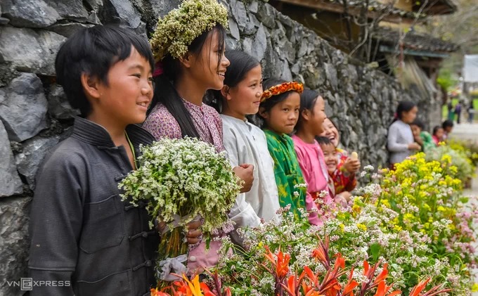 Trẻ em dân tộc Mông đứng bên những bức tường đá xếp trong bản để bán hoa cải, dong riềng, tam giác mạch... làm đạo cụ chụp ảnh cho khách. Mỗi bó hoa hoặc vòng đội đầu có giá 10.000 đồng.