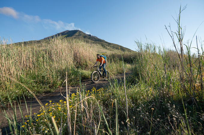 Du khách đạp xe khám phá núi lửa. Phía xa là miệng núi lửa đang hoạt động tỏa khói trắng lên bầu trời xanh. Con đường dẫn lên núi lửa thay đổi liên tục theo độ cao. Gần chân núi, con đường được lát bằng đá núi lửa từ năm 1951.
