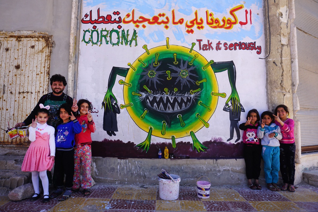  Nghệ sĩ người Syria, Aziz al-Asmar, cùng các trẻ em tạo dáng bên bức tranh minh họa virus SARS-Cov-2. Bức tranh được mô tả bằng tiếng Arab với chú thích: "Virus corona gây bệnh cho chúng ta. Hãy đối diện với nó một cách nghiêm túc".